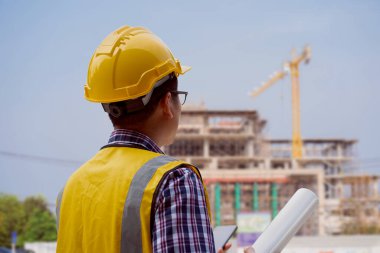 Sert şapkalı Asyalı bir erkek mühendis elinde bir plan tutuyor ve dışarıda dikilirken cep telefonuyla konuşuyor. Yapının yapısını izlemek ve kontrol etmek için ayakta durmanın boyutu