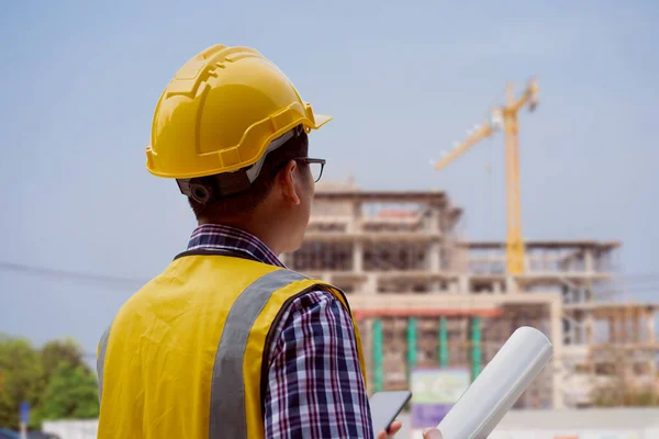 一位戴着硬礼帽的亚洲男性工程师一边站在室外 一边拿着一张蓝图 一边用手机交谈 监察及控制建筑物建造的立柱尺寸 — 图库照片