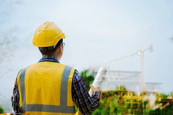 An Asian male engineer in a hard hat holds a blueprint and talks on his mobile phone while standing outdoors. Size of standing to watch and control the construction of the structure