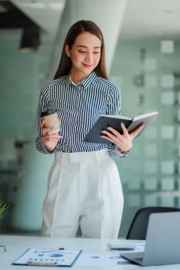 Happy successful Asian businesswoman holding a cup of coffee and tiger skin in front of her office desk.
