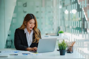 An Asian female accountant sits at a desk with a laptop and calculates financial graphs showing investment results. Plan a successful business growth process in the office.