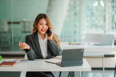 Asyalı bir kadın muhasebeci bir masada dizüstü bilgisayarla akıllı bir telefonla konuşuyor ve yatırım sonuçlarını gösteren bir mali grafik hesaplıyor. Ofiste başarılı bir iş büyüme süreci planlayın.
