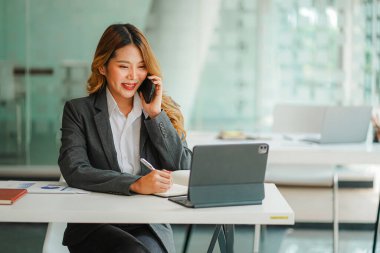 Asyalı bir kadın muhasebeci bir masada dizüstü bilgisayarla akıllı bir telefonla konuşuyor ve yatırım sonuçlarını gösteren bir mali grafik hesaplıyor. Ofiste başarılı bir iş büyüme süreci planlayın.
