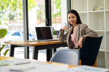 Asyalı iş kadını masasının önünde oturuyor kameraya bakıyor ve işini bitirirken mutlu bir şekilde gülümsüyor..