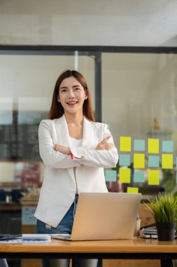 A happy Asian woman in a white dress stands at his desk smirking gleefully at a financially successful business.