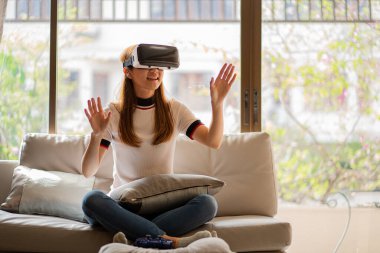 Neşeli beyaz kadın evde VR gözlük takıyor. Gözlüklerle eğleniyor. Elleriyle hayali jestler yapıyor..