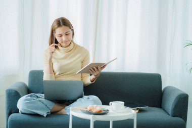 Kanepede oturan Asyalı kadın ev konseptinde dizüstü bilgisayarla çalışıyor.