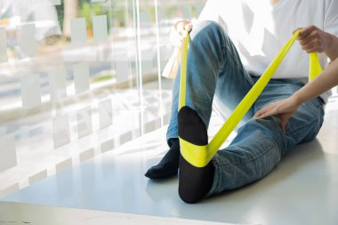 Rehabilitasyon terapisi. Bir fizyoterapist, yeşil-sarı bir hortumla bacağını esnetirken bir hastaya yardım eder ve rehabilitasyon merkezinde genç bir erkek hastayla çalışır..