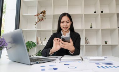 Ofiste resmi takım elbiseli Asyalı bir iş kadını ve hesap makinesiyle finansal grafikleri analiz etmek için bilgisayarla akıllı telefon kullanıyor..