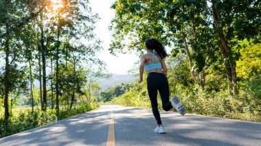 Spor kıyafetli ince bir kız aydınlık bir sabahta yolda koşuyor, sonbahar ormanında koşuyor. Arkadan bak. Yoldayım. Açık havada çekim..