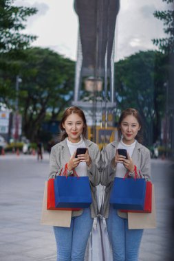 Çanta tutan Asyalı kız alışveriş merkezinde alışveriş yaparken akıllı telefonunu kullan. Alışveriş merkezinin önündeki festivalde alışveriş yaptıktan sonra bolca alışveriş yapan güzel bir kızın portresi..