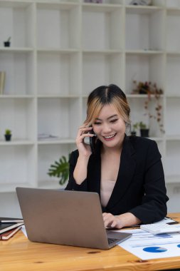 Asyalı bir kadın ofis masasında bilgisayar başında çalışırken akıllı telefon ya da cep telefonuyla konuşur..