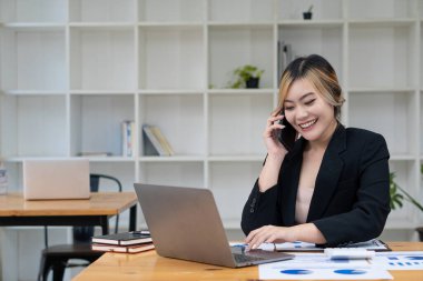 An Asian woman talks on a smartphone or mobile phone while working on a computer at her office desk.