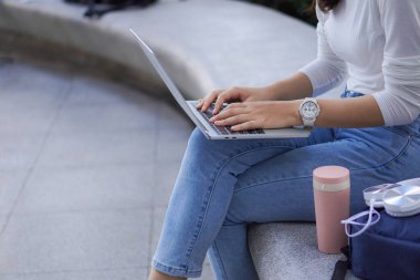 Genç bir kadın yerde oturup dizüstü bilgisayarda klavye yazıyor. İnternette kulaklıklar ve açık havada çalışma fikrinin yanında bir bardak suyla çalışıyor..