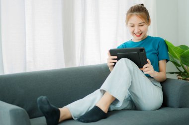 Yatak odasındaki kanepede tablet kullanan güzel bir Asyalı kadının portresi. Mutlu kadın evde kanepede oturup arkadaşlarıyla film ve video izlemek için tablet kullanıyor..