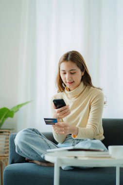 Güzel Asyalı kadın internet sitesinden alışveriş yapıyor ve kredi kartını ödeme için kullanıyor. İnternetten alışveriş yapmak, kanepede oturmak.