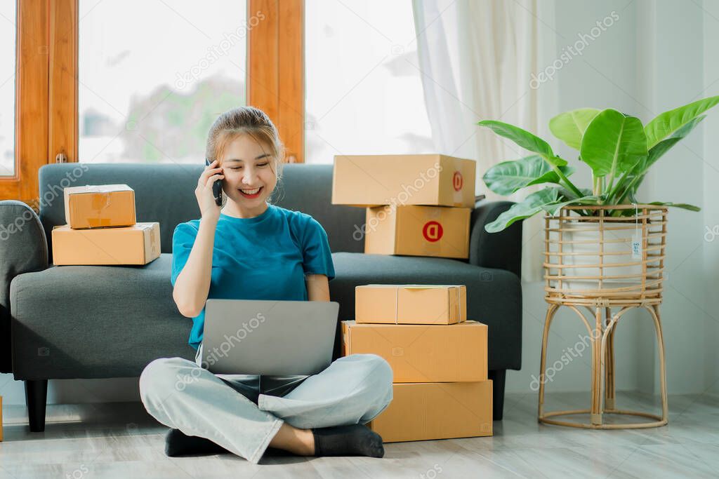 A young Asian woman starts a business to talk to customers via smartphone. Work happily. with box and laptop at home Prepare to deliver parcels in SMEs, supply chains, online procurement concepts.