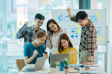 Genç Asyalı iş grupları mutlu ve başarılı hissediyorlar. İş hedeflerindeki başarıları kutlamak için gülen eller masadaki dizüstü bilgisayarla ve ofisteki finans grafiğiyle uğraşır.