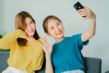 İki Asyalı kadın akıllı telefonlarıyla gri koltuklarda oturup evlerinde sosyal medyada fotoğraf çekiyorlar..