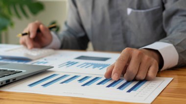 İş adamı ellerinin kalem tutuşu pazarlama grafiğine işaret ediyor ve bilgisayarda ve finansal belgelerde çalışıyor. Ofiste online muhasebe işi kavramı