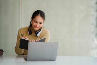 Boynunda kulaklık olan çekici ve mutlu Asyalı kız öğrenci oturma odasındaki masada oturuyor gülümsüyor ve iş yerinde dizüstü bilgisayar, defter, genç girişimci ya da serbest meslek fikri üzerine çalışıyor..