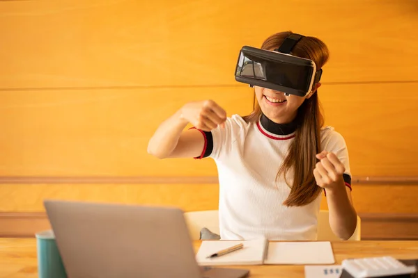 Cheerful caucasian woman wearing VR glasses at home. Young woman enjoying glasses with imaginary hands doing imaginary gestures on virtual reality device at desk with laptop and calculator documents.
