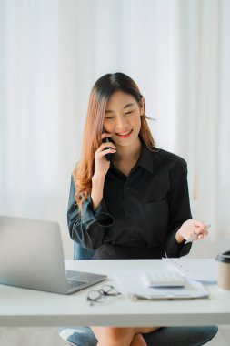 Akıllı telefon tutan Asyalı iş kadını, elinde kalem, dizüstü bilgisayar ve hesap makinesiyle grafiği gösteren bir raporu analiz ediyor. iş istasyonu muhasebecisinde çevrimiçi pazarlama konsepti e-ticaret dikey imajı