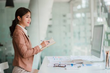 Asyalı iş kadını, grafiği gösteren bir raporu kalem, dizüstü bilgisayar ve hesap makinesiyle analiz ediyor. İş istasyonu muhasebecisinde çevrimiçi pazarlama konsepti eTicari manzara