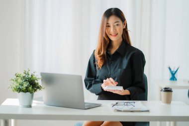 Asyalı iş kadını, grafiği gösteren bir raporu kalem, dizüstü bilgisayar ve hesap makinesiyle analiz ediyor. İş istasyonunda, muhasebede, çevrimiçi pazarlama konseptinde, e-ticarette, manzara resminde.