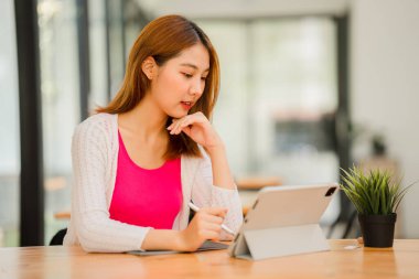 İnternette okuyan Asyalı kız öğrenci, tabletli e-öğrenim masasında oturuyor ve elinde kalemle mutlu Asyalı ilkokul öğrencisi kameraya gülümsüyor, uzaktan öğrenmeyi düşünüyor..