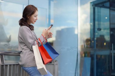 Alışveriş merkezinin zemininde alışveriş torbaları taşıyan gülümseyen Asyalı kadın. Kadın modası, alışveriş, yaşam tarzı, mutluluk, tüketim, satış ve insan kavramı.
