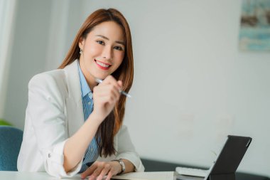 Asyalı bir kadın, elinde tabletle modern bir ofiste masa başında oturuyor ve müşterilerle mesajlaşarak internet üzerinden sohbet ediyor..