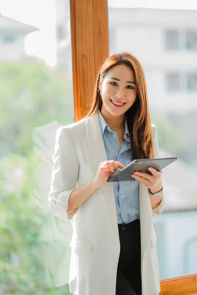 A corporate executive, a casual Asian businesswoman, uses a digital tablet in her home workspace to connect with customers through an office lifestyle adapted to online technology.