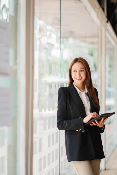 A corporate executive, a casual Asian businesswoman, uses a digital tablet in her home workspace to connect with customers through an office lifestyle adapted to online technology.