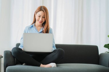 Kanepede dizüstü bilgisayarla oturan Asyalı kadın oturma odasında lüks bir kanepenin keyfini çıkarıyor..