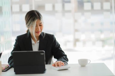 Asyalı bir kadın, elinde tabletle modern bir ofiste masa başında oturuyor ve müşterilerle mesajlaşarak internet üzerinden sohbet ediyor..