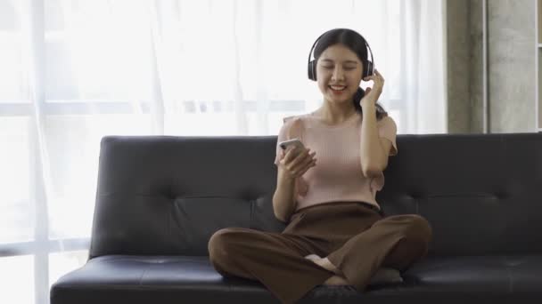 ヘッドフォンをしている若いアジアの女性は 自宅のソファで音楽を喜んで聞くことができます — ストック動画