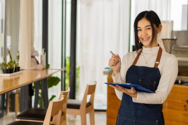 Önlüklü güzel Asyalı barista elinde tabletle dükkan sahiplerinin, girişimcilerin, KME girişimcilerinin, konseptlerin bir kahve tezgahının önünde duruyor..