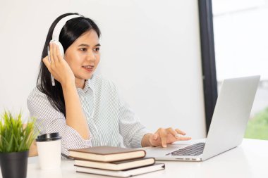Bağımsız iş kadını kulaklık takıyor, bilgisayarı kullanıyor, iş yerini kullanıyor, iş yerindeki müşterilerle iş yerinde video görüşmesi yapıyor. Mutlu Asyalı genç kız İnternet 'te rahatlıyor..
