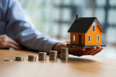 Savings accountant holding a small wooden house in hand and pile of coins on the table, financial and accounting concepts.