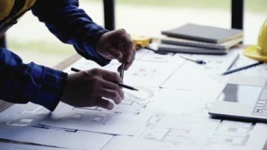 Mimari Proje Mühendisi inşaat projesini hazırlıyor, ev çizimlerini kağıt üzerinde kontrol ediyor, mimarlık ofisindeki masada dizüstü bilgisayar ve şapka ile tamir araçları için ev planı planlarını kontrol ediyor..