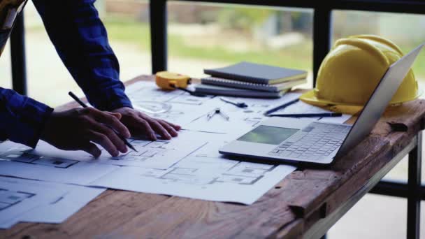 建築プロジェクト職場エンジニアは 建設プロジェクトを草案 紙の上の家の図面をチェックし ラップトップと帽子で建築事務所の机の上の修理ツールのための家の計画設計図 — ストック動画
