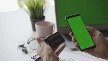 Bir adamın elinde kredi kartı var, evdeki modern akıllı telefon ya da cep telefonunu kullanarak internetten ödüyor. Masada oturmak, finans, internet alışverişi.