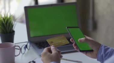 Bir adamın elinde kredi kartı var, evdeki modern akıllı telefon ya da cep telefonunu kullanarak internetten ödüyor. Masada oturmak, finans, internet alışverişi.