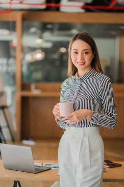 Asyalı iş kadını elinde bir fincan kahve ile yatırım projeleri ve finansal muhasebe planlama stratejileri üzerinde çalışıyor. İşadamı ofiste dizüstü bilgisayarla konuşuyor.