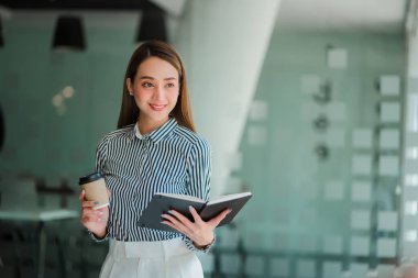 Asyalı iş kadını elinde bir fincan kahve ile yatırım projeleri ve finansal muhasebe planlama stratejileri üzerinde çalışıyor. İşadamı ofiste dizüstü bilgisayarla konuşuyor.