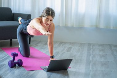 Gülümseyen Asyalı kadın evinde dizüstü bilgisayardan yoga yaparak omuz geriyor. Yanında bir kanepe ve dizüstü bilgisayarla evde yalnızlık ve egzersiz..