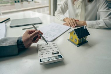 Emlakçı ev satın alma sözleşmesinin şartlarını tartışır ve müşteriden sözleşmeye yasal olarak girmek için belgeleri imzalamasını ister. Ofis masasında ev satmak için fikirler.