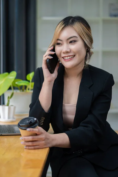 good business talk Beautiful Asian woman talking on mobile phone and playing laptop with smile while sitting at her desk. Share good business stories