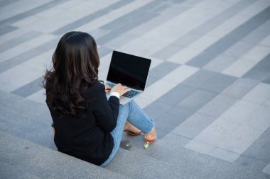 Diz üstü bilgisayarı olan Asyalı bir iş kadını iş bölgesindeki ekranlara bakıyor, profesyonel kadınlar dışarıda oturuyor.